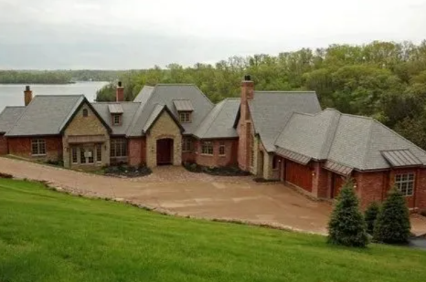 Large brick house with gray roof on a green hillside, surrounded by trees and a paved driveway
