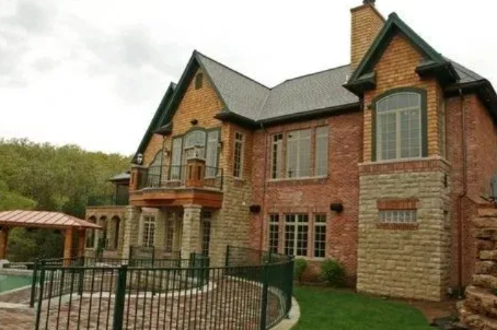 Brick two-story house with a balcony, large windows, and a fenced patio in a wooded setting
