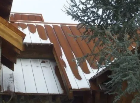 Snow-covered brown metal roof with patches of white snow beside a tree