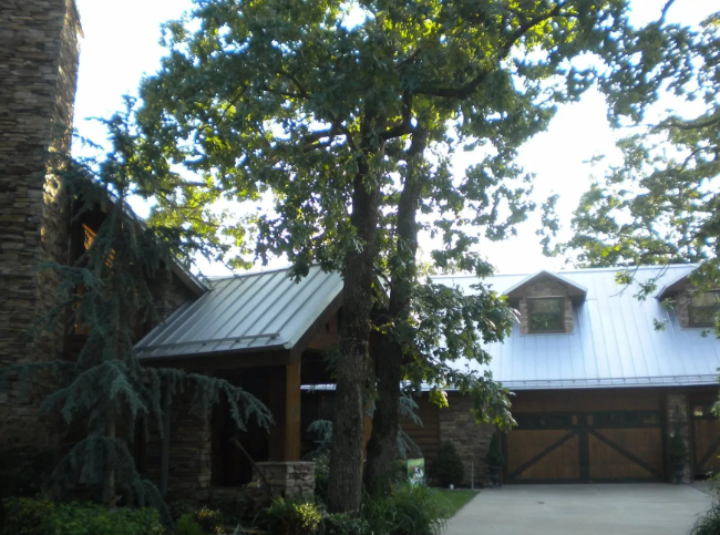 Stone house with a metal roof, large trees, and a garage door in a wooded setting