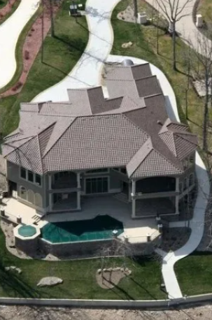 Aerial view of a large house with a gray roof, backyard pool, and curved driveway, surrounded by lawn and trees