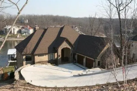 Large tan house with dark roof and curved driveway in a wooded suburban setting