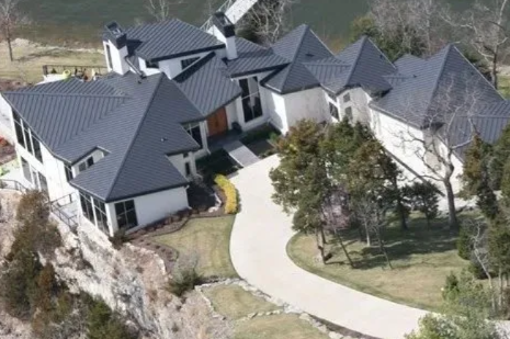 Aerial view of a large house with dark gray roofs and a curved driveway surrounded by trees and lawn