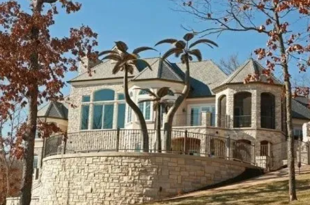 Large stone house with a curved balcony, palm trees, and autumn trees on a sunny day