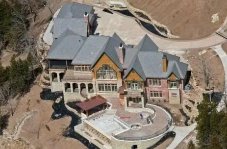 Aerial view of a large beige mansion with gray roofs and a backyard pool on a wooded hillside