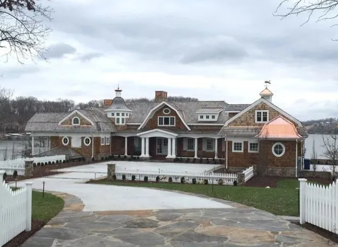 Large brown shingle house with white trim and a circular driveway under a cloudy sky