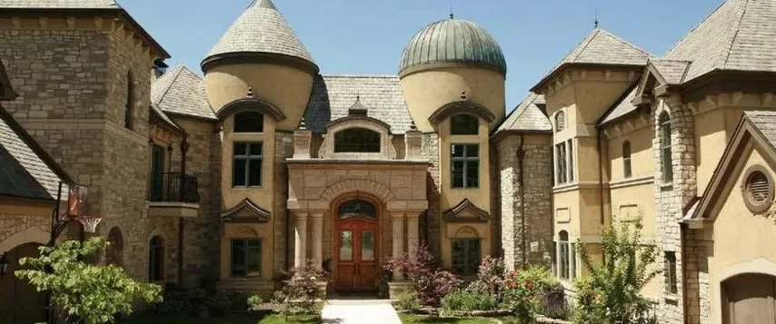 Stone mansion with domed roofs, arched windows, and a central wooden entrance surrounded by greenery