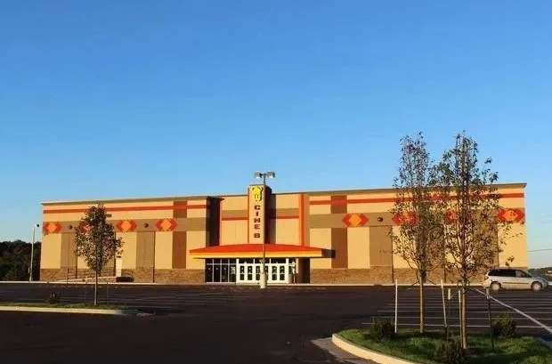 Large beige and orange building with a central entrance, trees, and a parking lot under clear sky