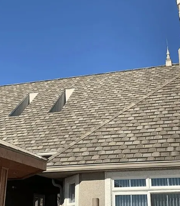House roof with gray shingles, two skylights, and a blue sky above a windowed exterior wall