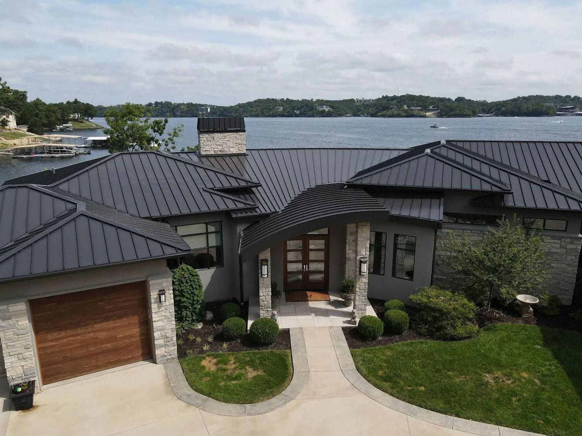 Modern waterfront house with dark metal roof, stone facade, and curved driveway