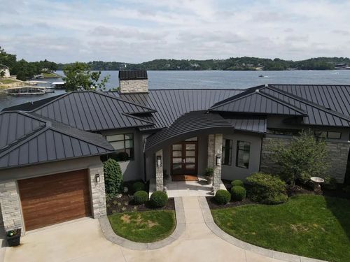 Modern waterfront house with dark metal roof, stone facade, and curved driveway.