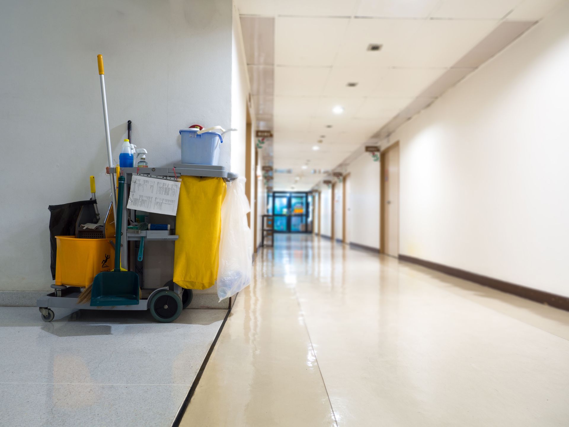 Janitor's cleaning cart in a bright, empty hallway with glossy floor.