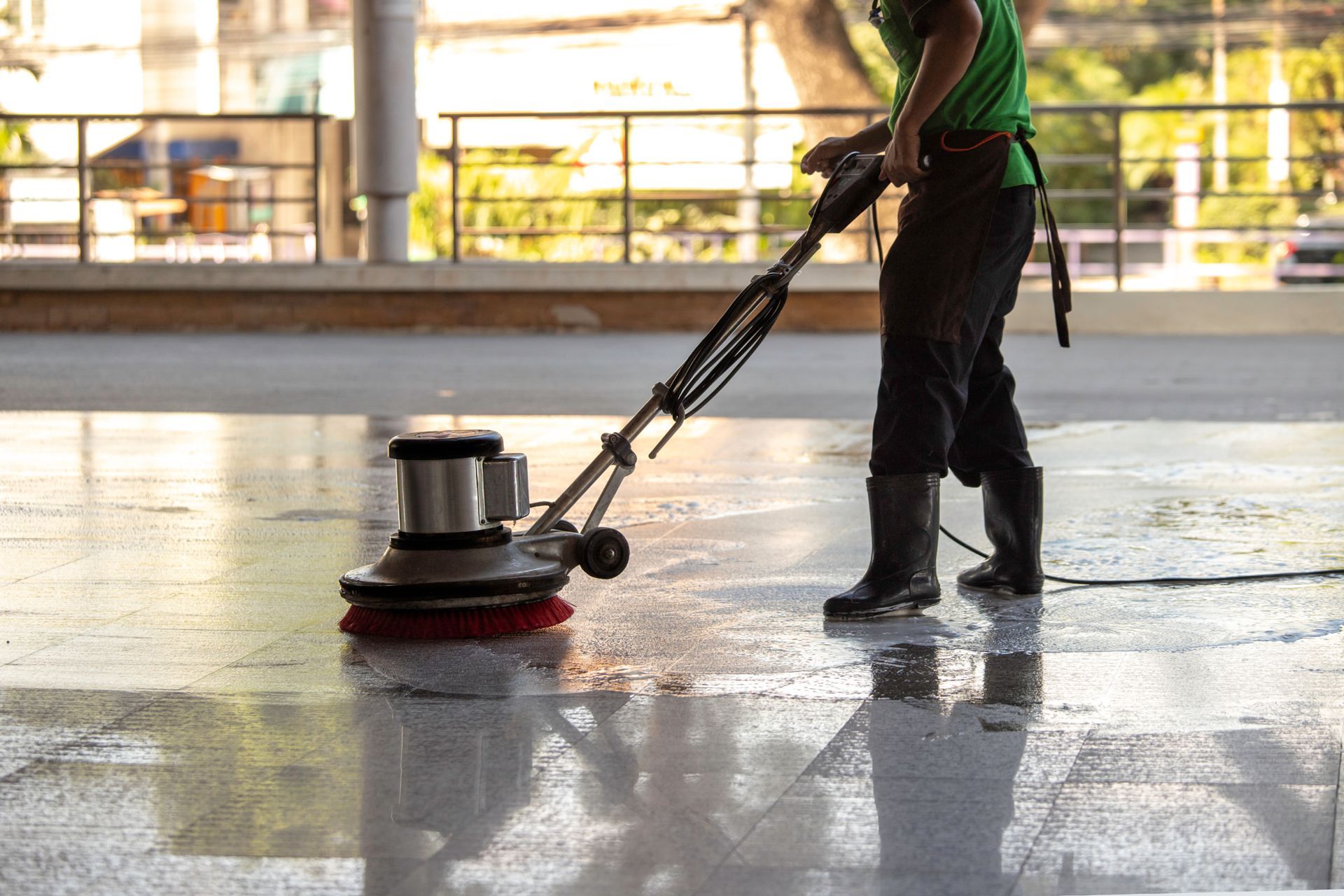 Man using a floor buffer to clean a glossy concrete floor outdoors, wearing green shirt and black pants.