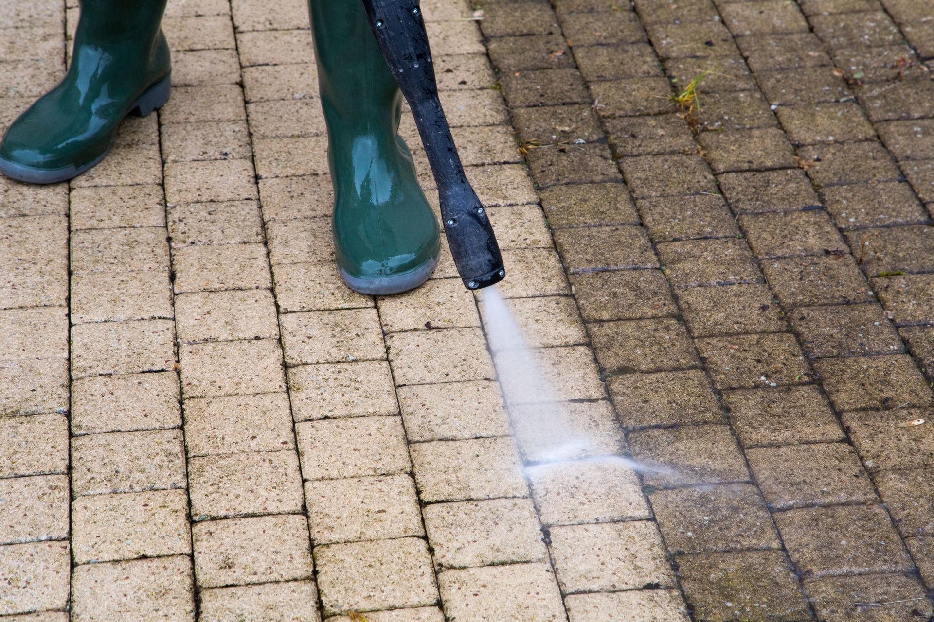 Person in green boots using a power washer on brick pavers, showing a clear cleaning effect.