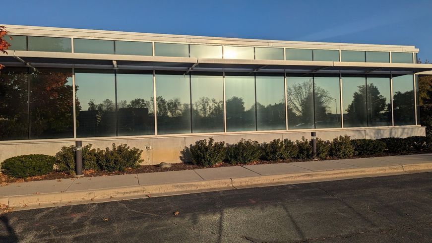 Empty glass-fronted storefront with a flat roof and shrubs along the base, viewed from a parking lot.