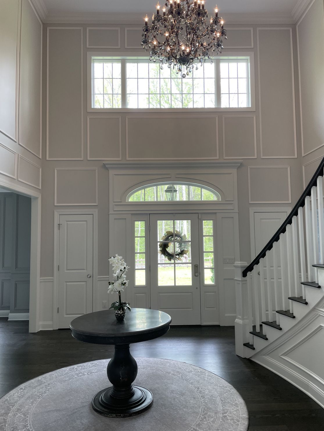 Elegant foyer: white trim, chandelier, dark wood table with floral arrangement, staircase, front door with wreath.