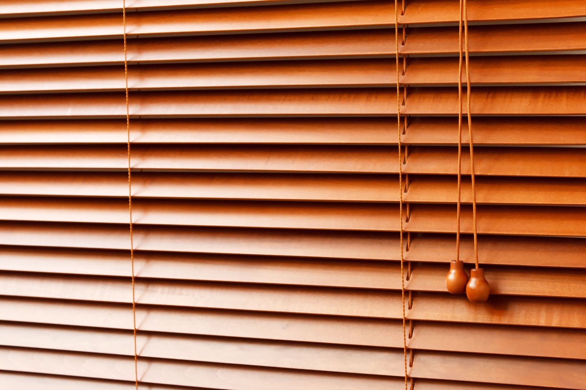 Closed wooden window blinds in warm brown tones with hanging pull cords.