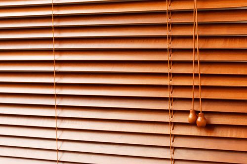 Closed wooden window blinds in warm brown tones with hanging pull cords.