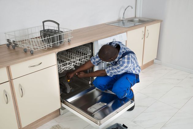 A person in blue overalls fixing a dishwasher in a kitchen.