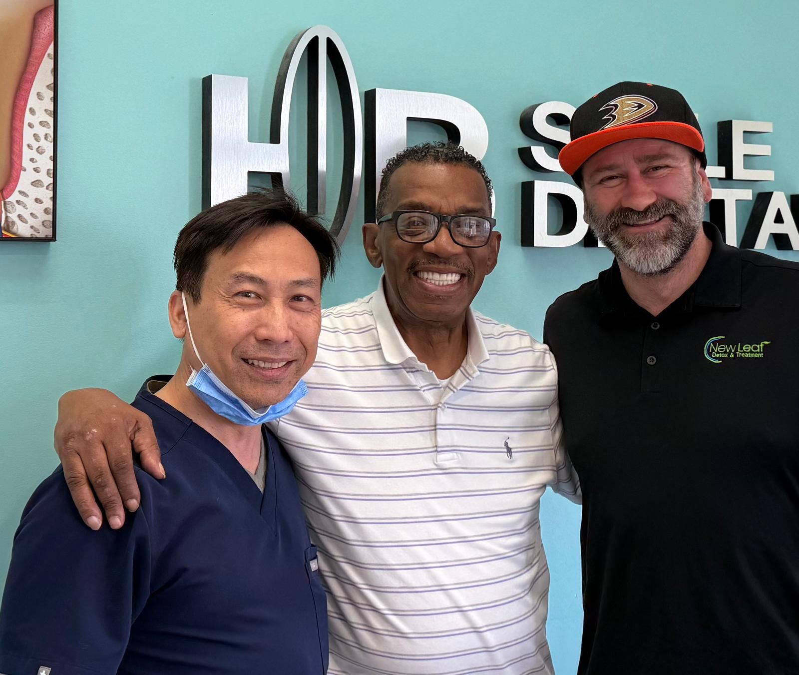Three smiling people posing in a dental office: a patient with two dental professionals.