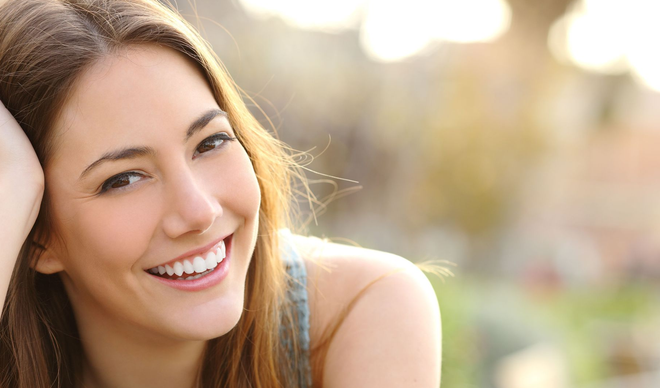 Woman smiling, outdoors; brown hair, blue top, soft sunlight in the background.