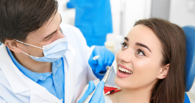 Dentist examining a patient's teeth. The woman is smiling, in a dental chair. Blue gloves and a mask are worn.