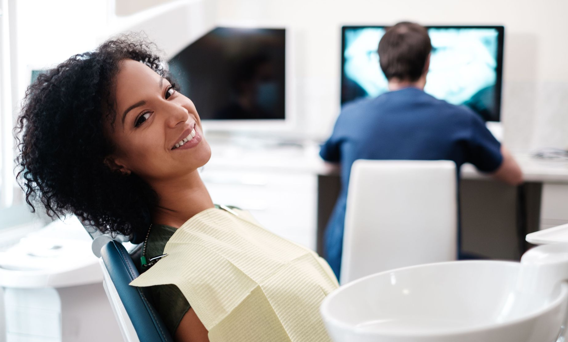 Woman smiles in a dentist's chair; dental professional in background.