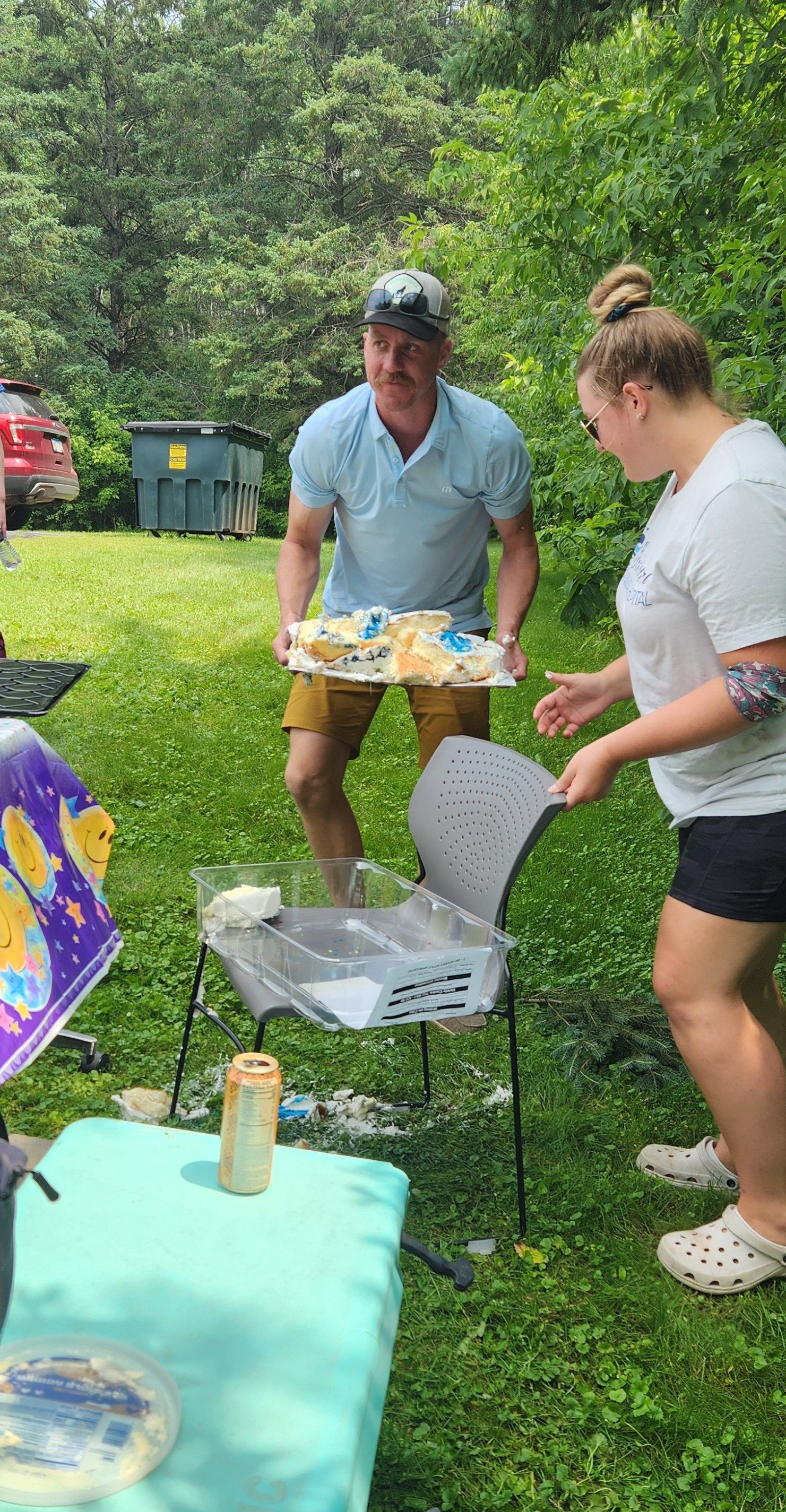 Man carrying cake outdoors, being assisted by a woman. They stand near a table and chair in a grassy area.