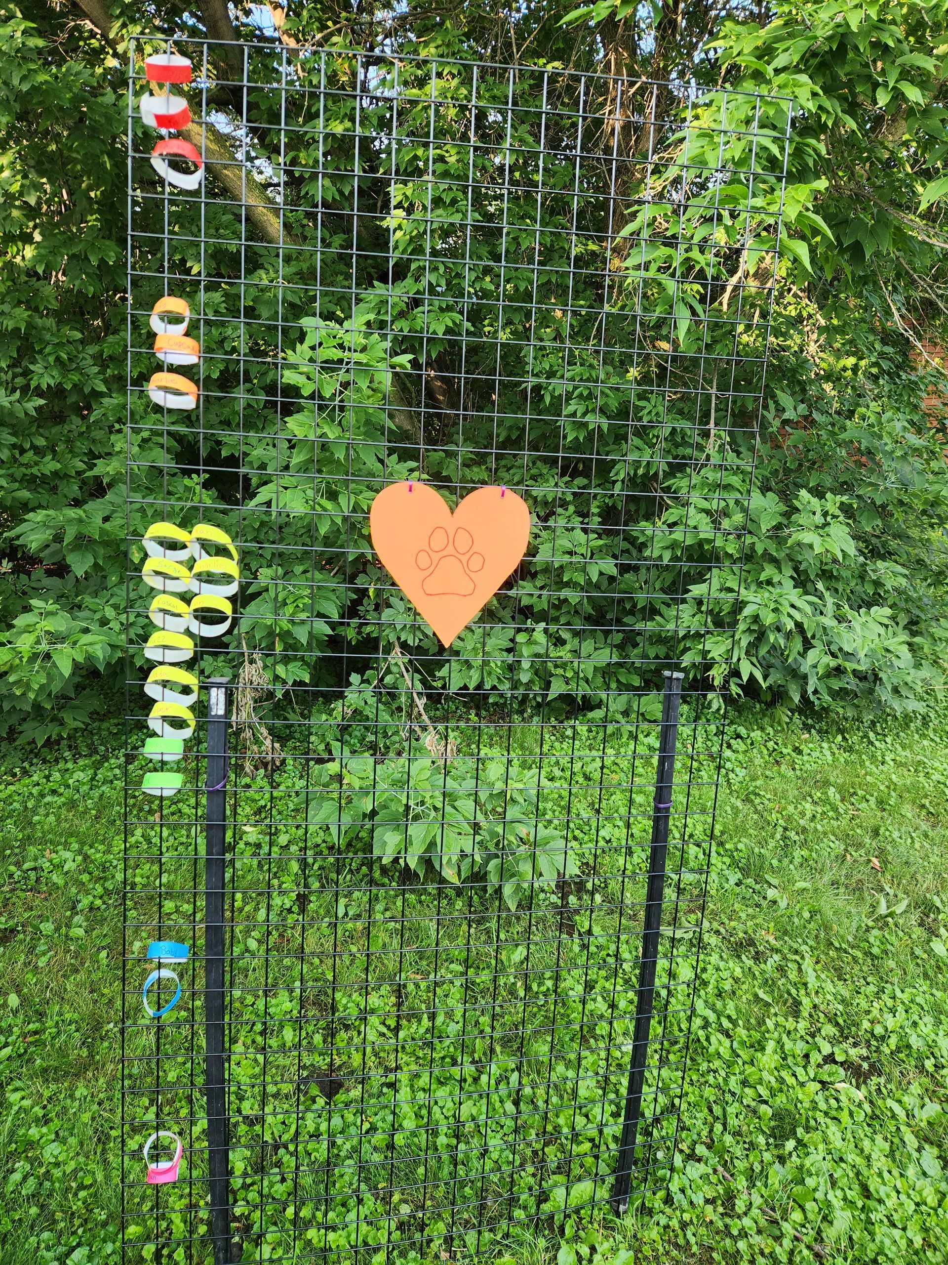 A metal art installation with hanging objects and an orange heart against a backdrop of green trees and grass.
