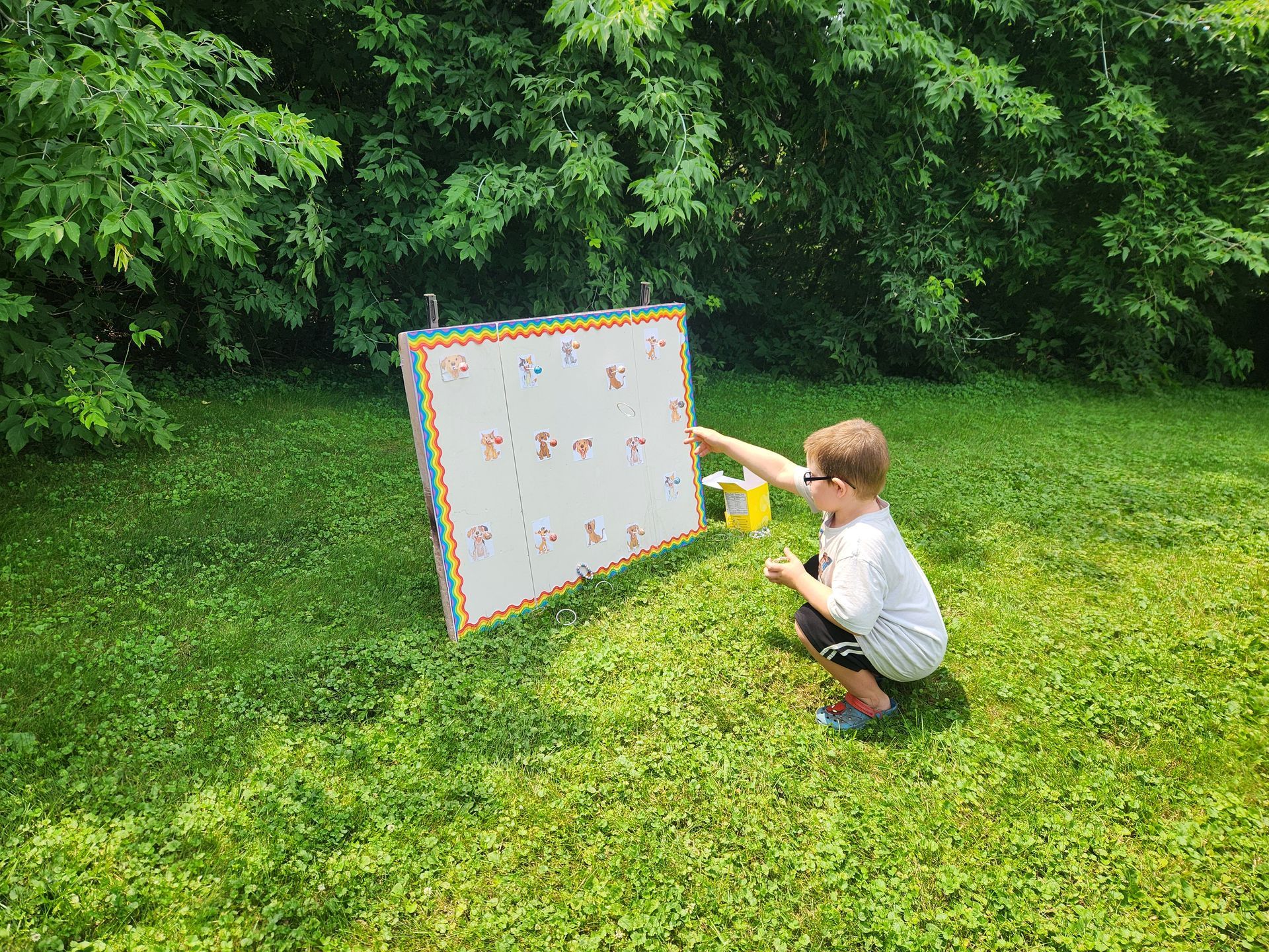 A young boy, wearing glasses, squirts a yellow substance onto a white board with colorful markings in a grassy outdoor setting.