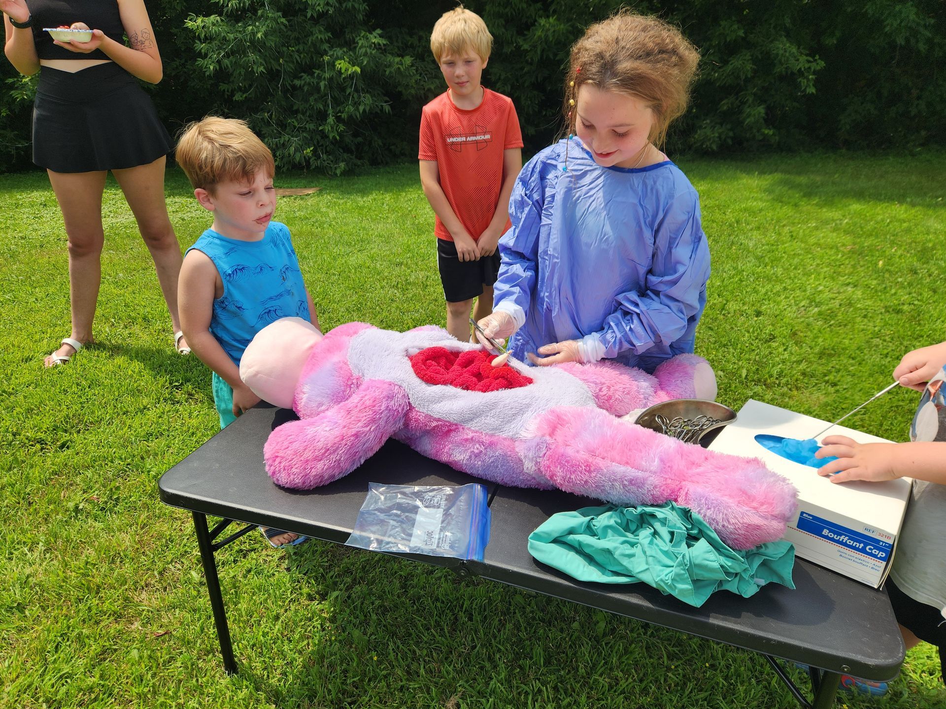 Children perform a pretend surgery on a stuffed animal outside. One child in a surgical gown uses a tool, others watch.
