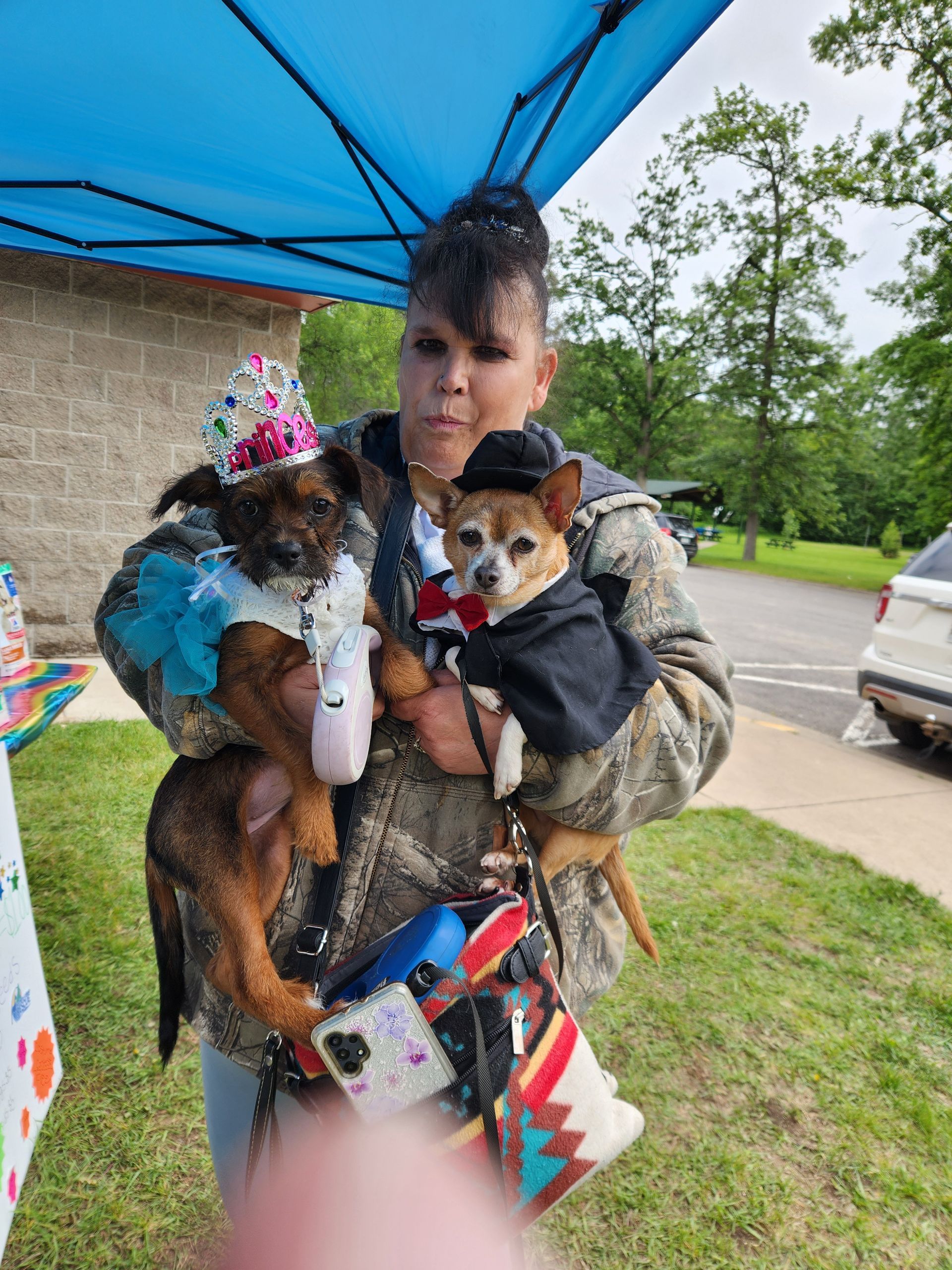 Woman holding two small dogs in costumes. One wears a hat with a bow, the other, a tuxedo. Outdoors.