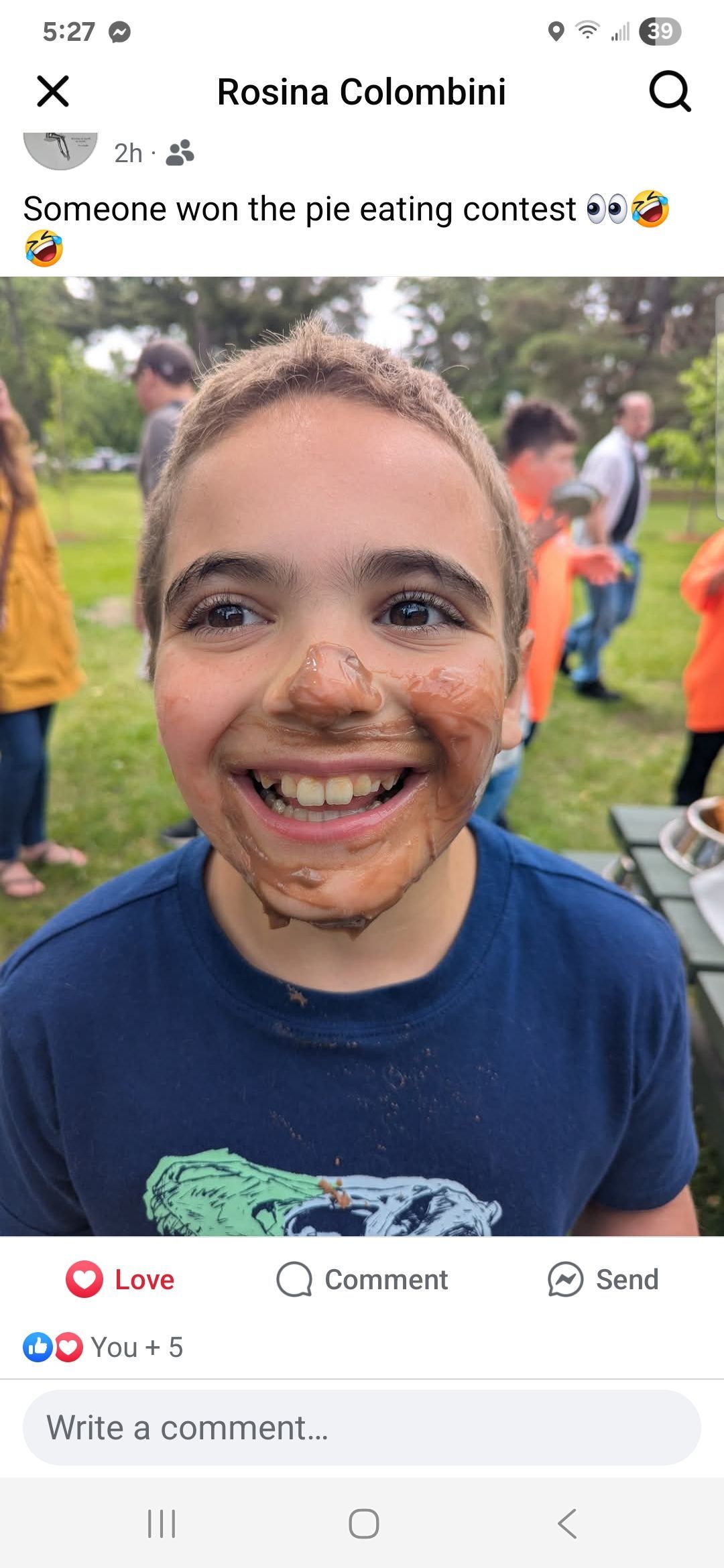 Boy with muddy face smiling, outdoors with people in the background.