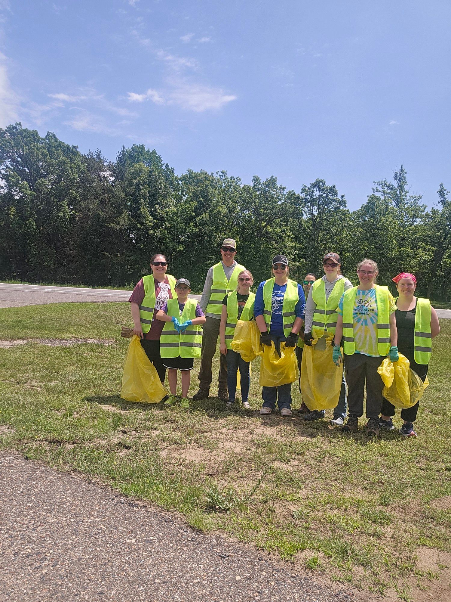 Group of people in safety vests collecting trash in a grassy area on a sunny day.
