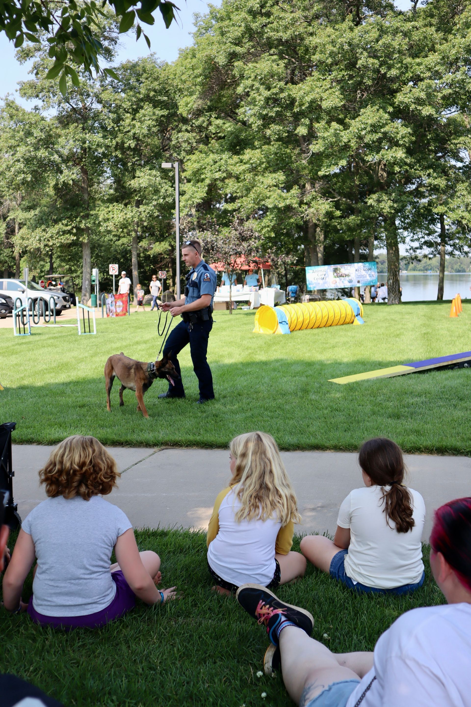 A group of people are sitting on the grass watching a police officer train a dog.