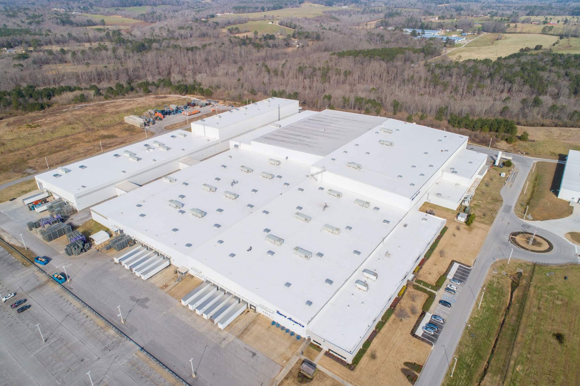 Aerial view of a large white warehouse building with loading docks and surrounding parking lot; trees and hills in the background.