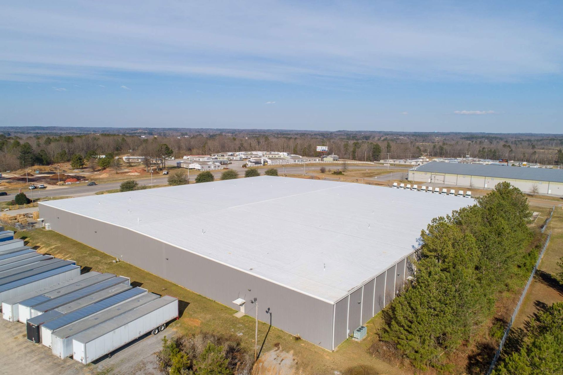 Large industrial building with a white roof and metal siding, surrounded by trees and other buildings.