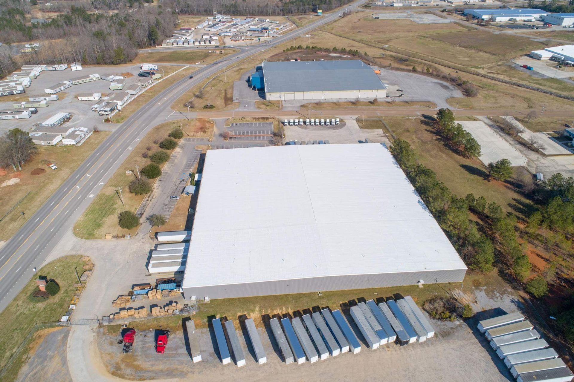 Aerial view of a large industrial building with a white roof, surrounded by trailers, vehicles, and other buildings.