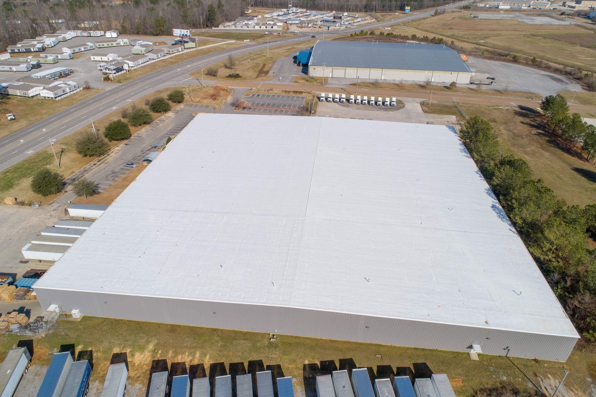 Aerial view of a large industrial building with a white roof, next to a road and other buildings.