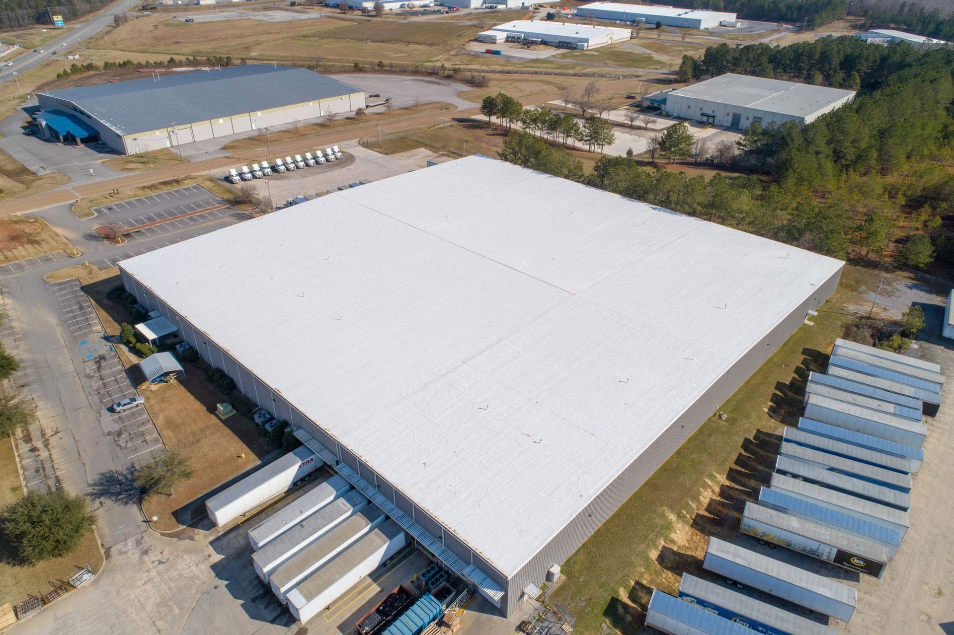 Aerial view of a large warehouse with a white roof, surrounded by other buildings and parked semi-truck trailers.