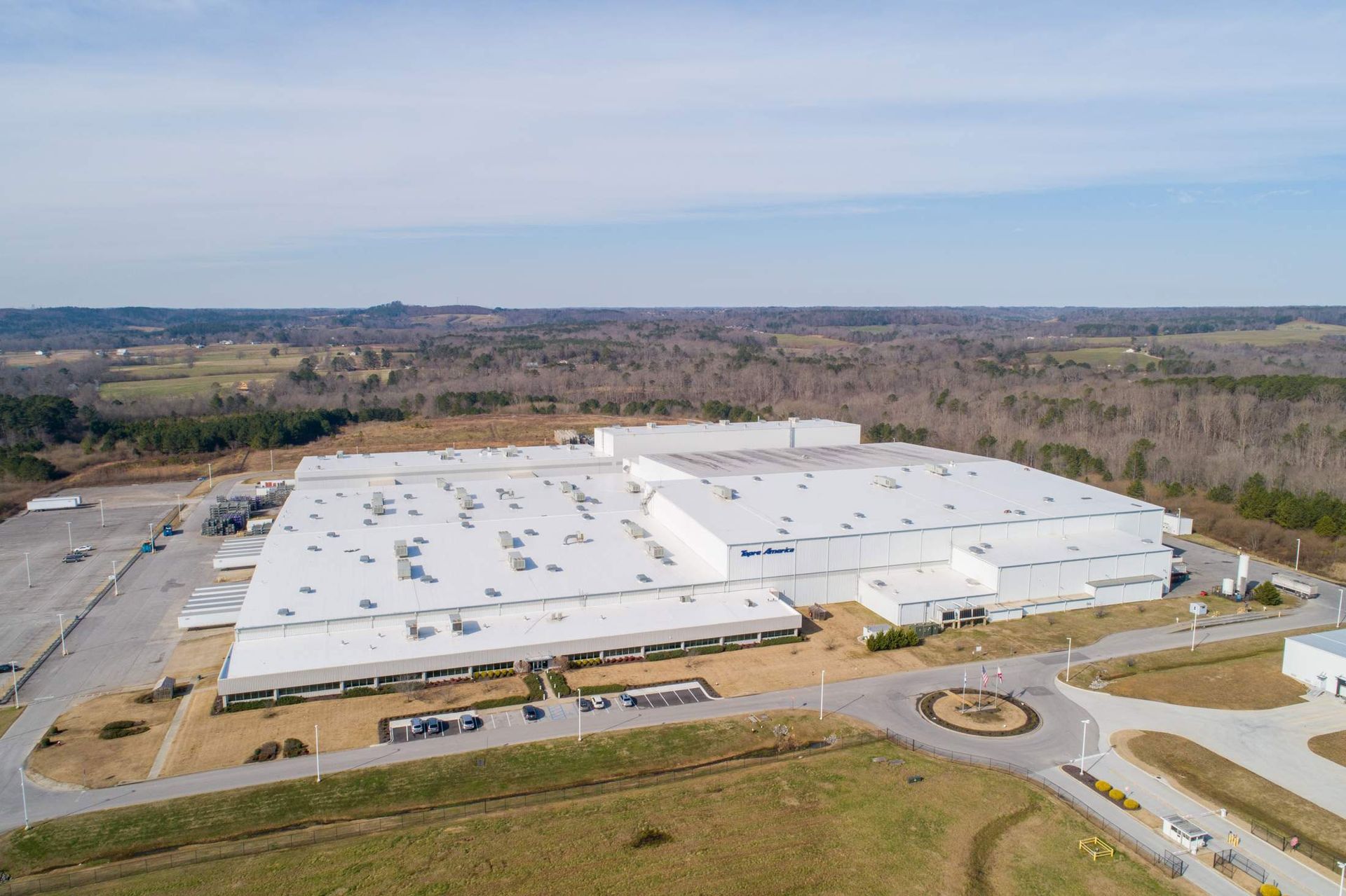 Aerial view of a large industrial building with a white roof surrounded by a parking lot and trees under a blue sky.