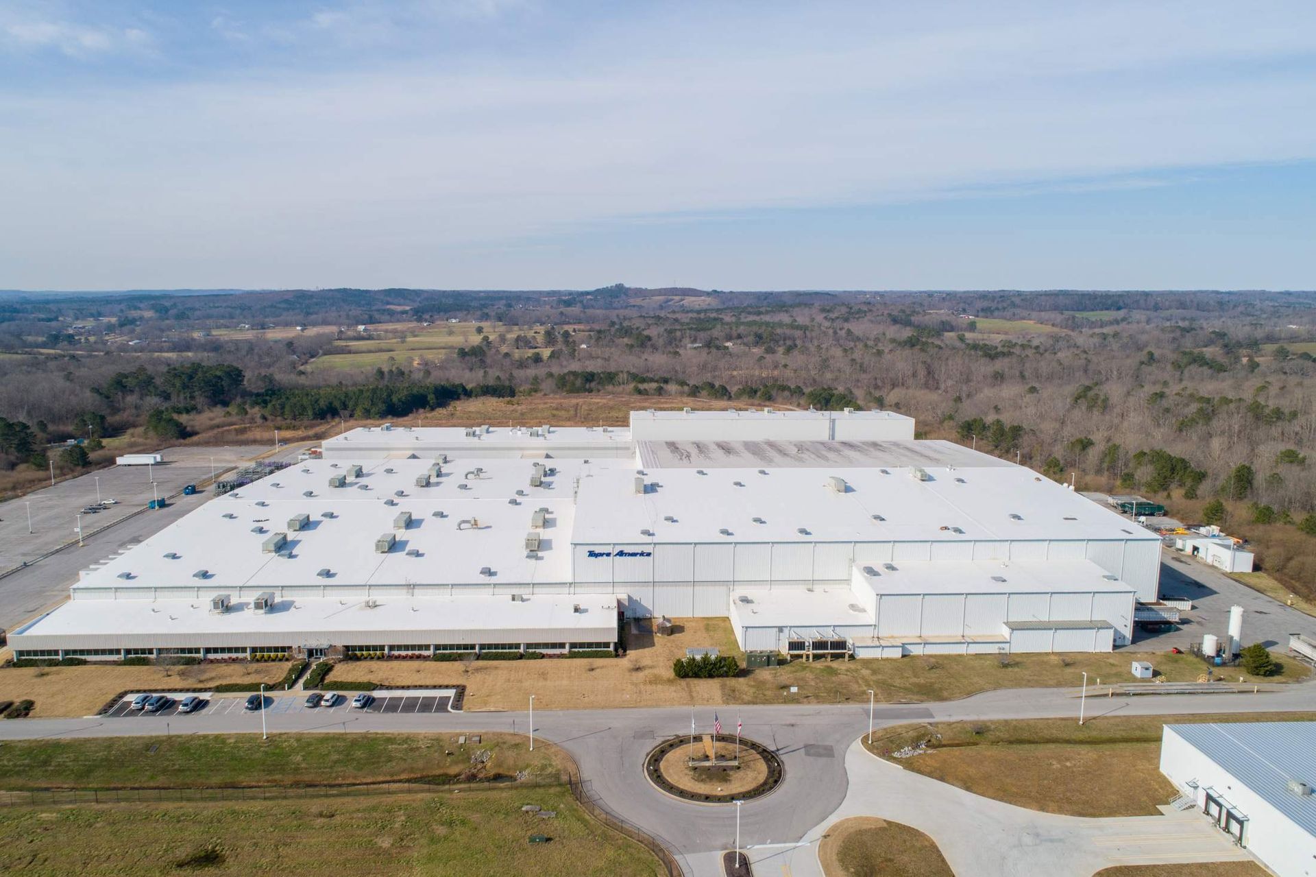Aerial view of a large, white industrial factory building with a circular driveway in front.
