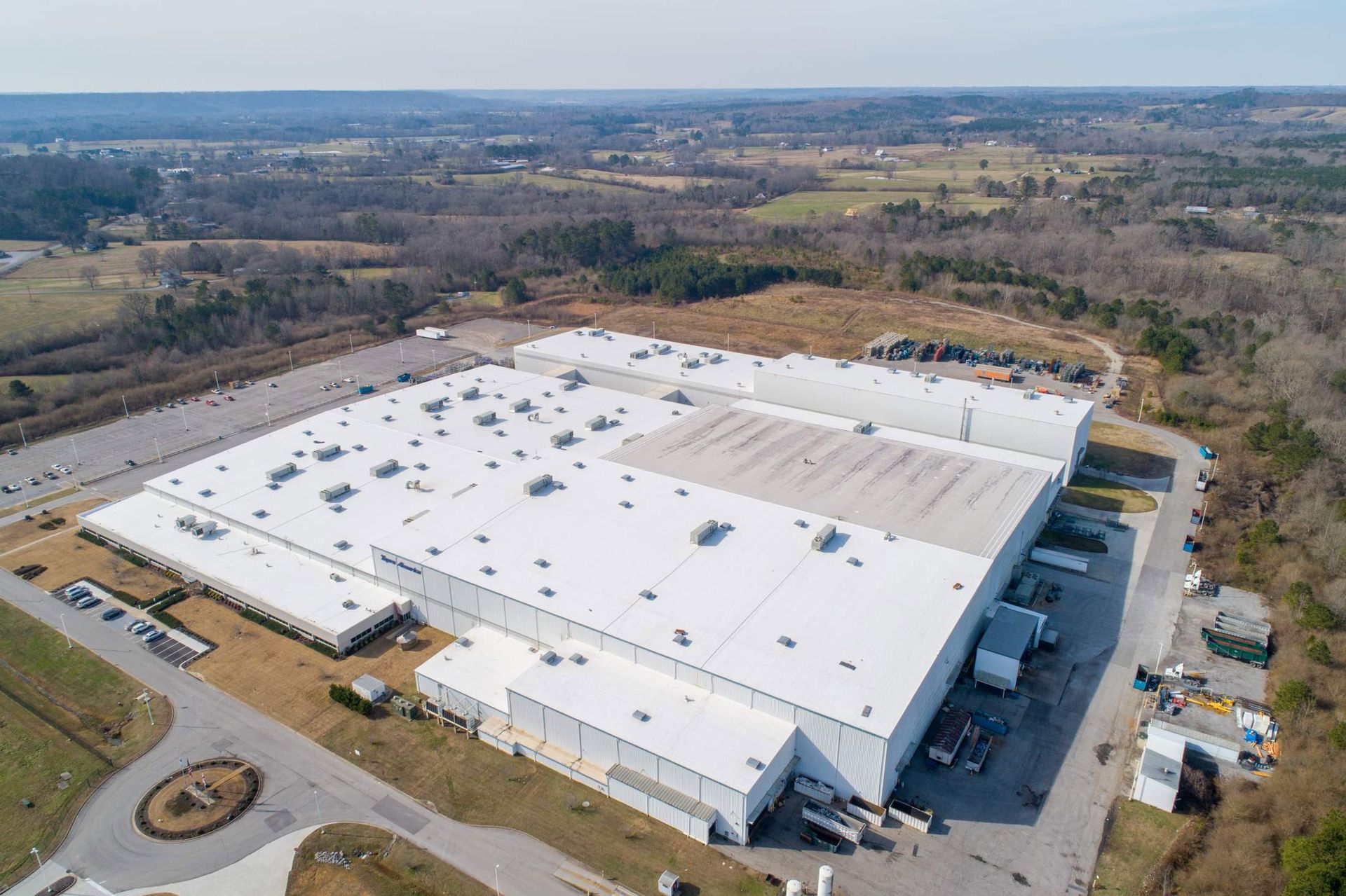 Aerial view of a large white industrial building with a parking lot in a rural landscape.