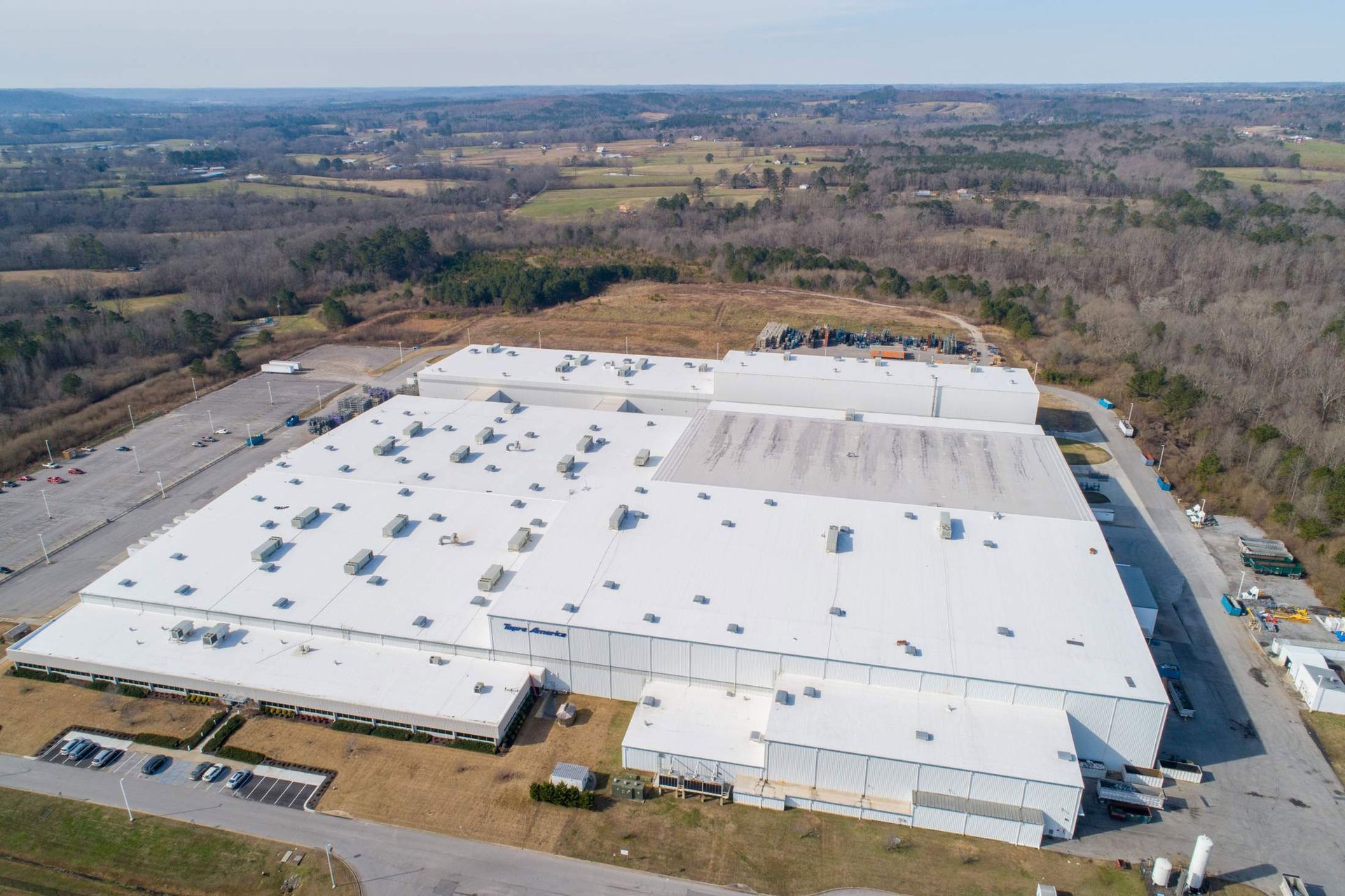 Aerial view of a large, white-roofed factory building surrounded by a parking lot and fields.