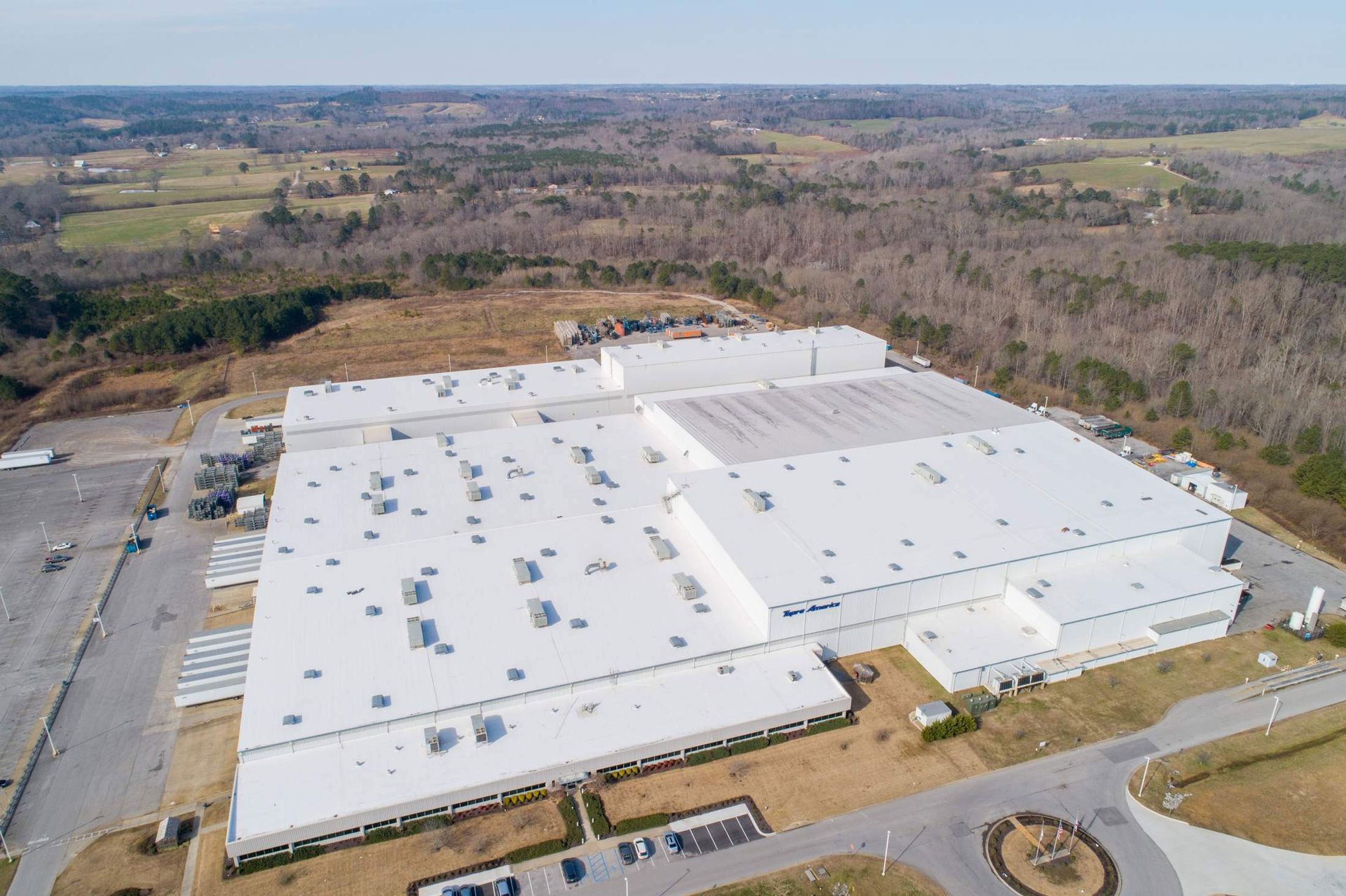 Aerial view of a large white industrial building with a flat roof, surrounded by trees and greenery.
