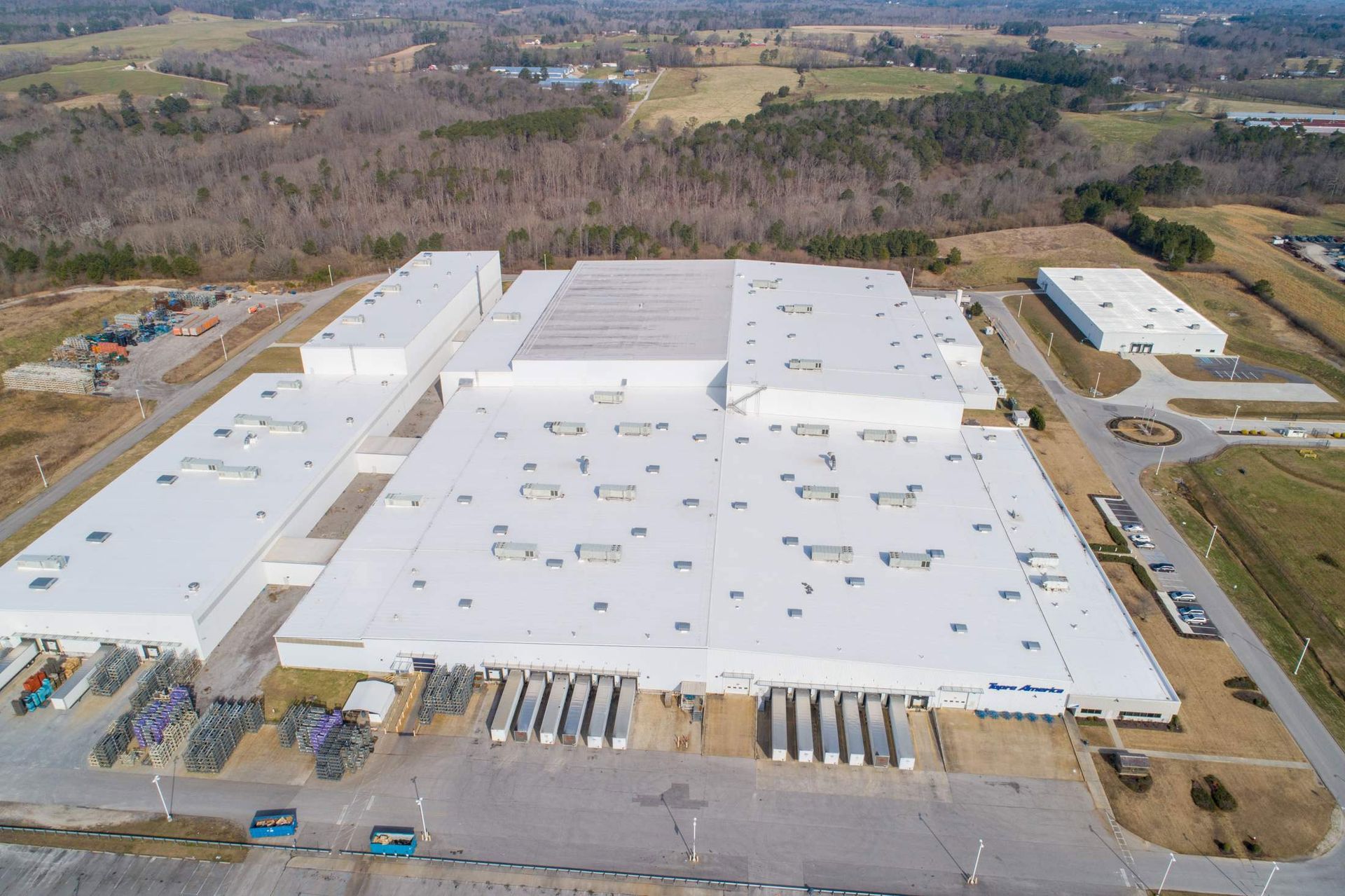 Aerial view of a large white industrial building with loading docks and a parking lot. Trees and fields in the background.