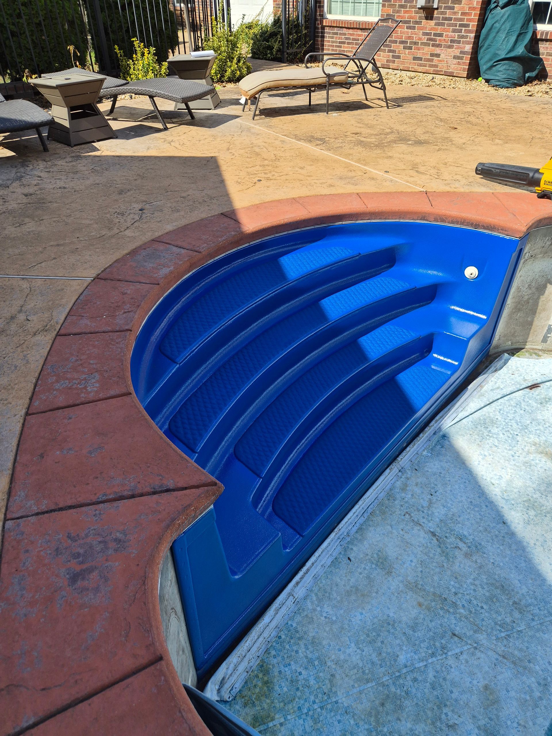 Blue pool steps with a red-brown border, partially submerged in a pool. Concrete patio in background.