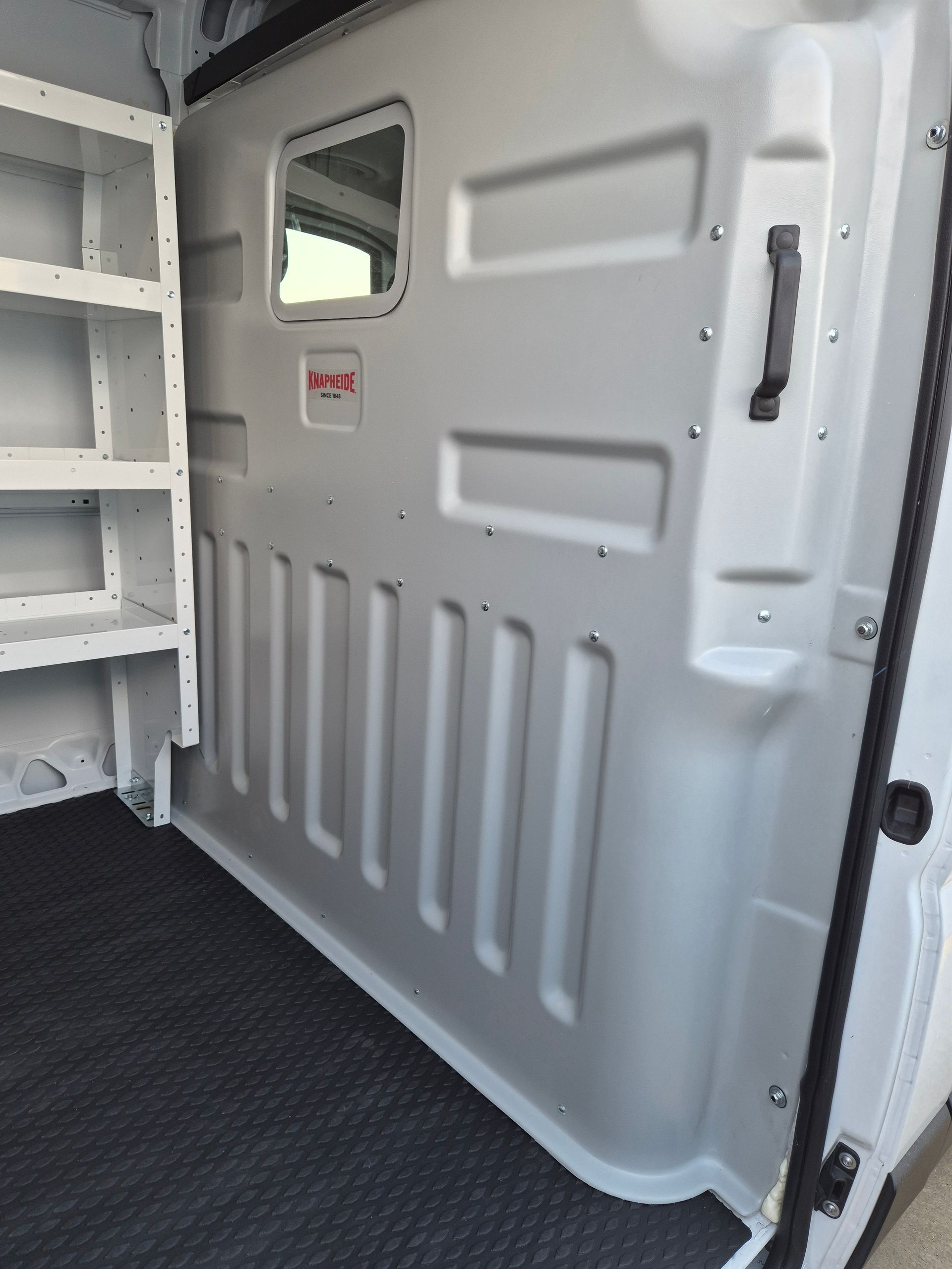 Interior of a cargo van with shelving on the left and a wall with a window and handle on the right.