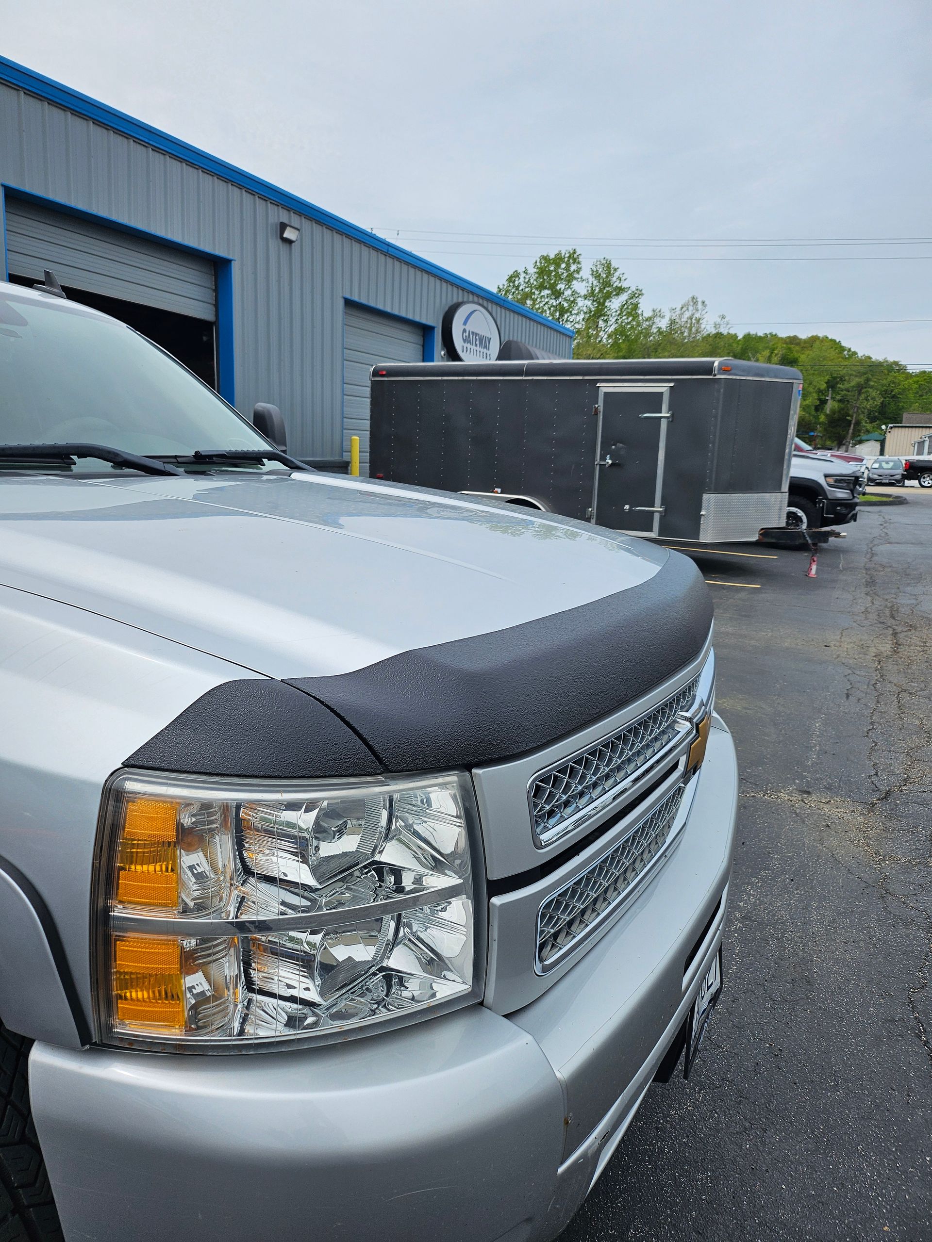 Silver pickup truck parked in front of a building with a trailer. The hood has a black bug deflector.