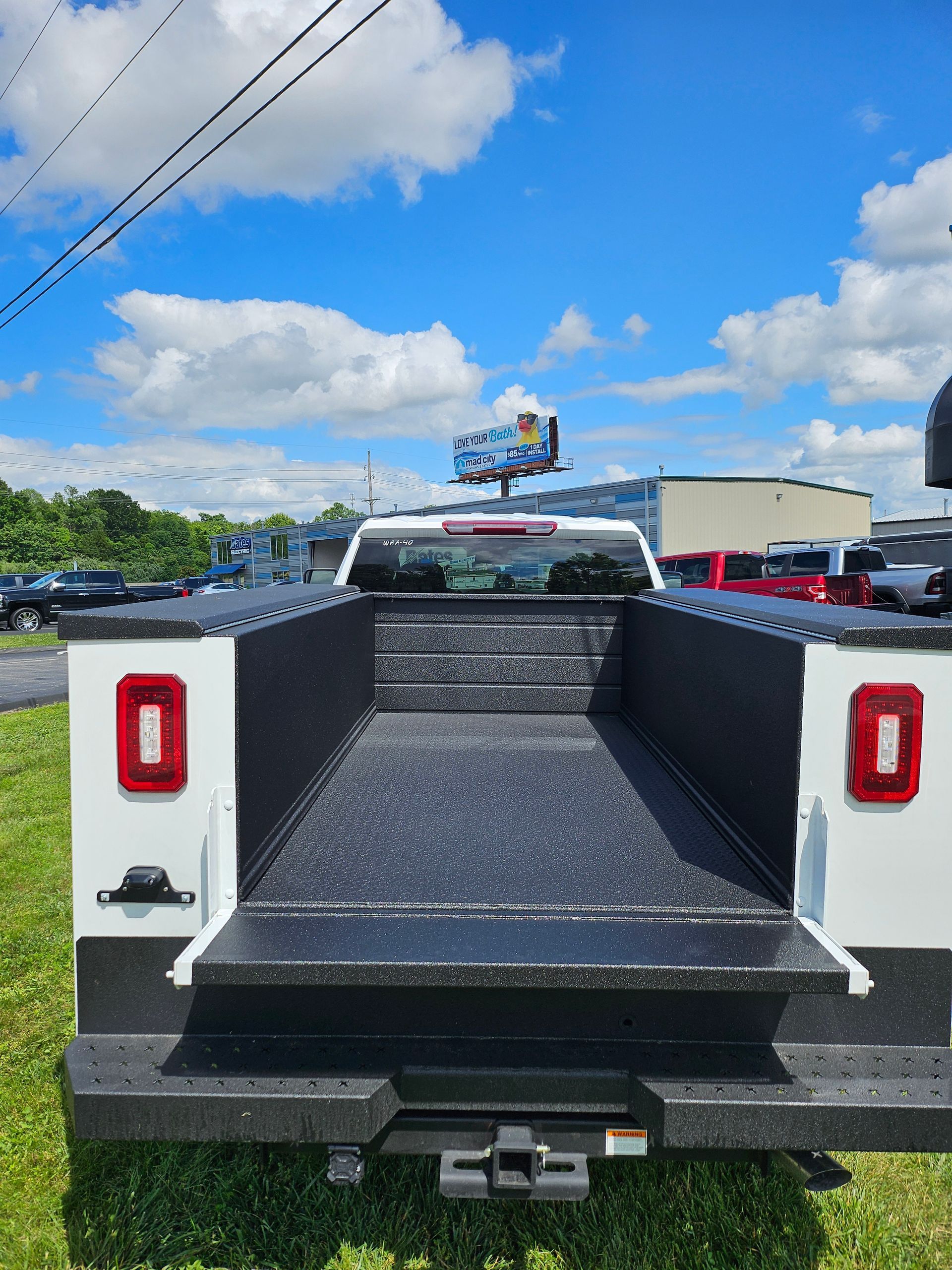 White work truck bed with black liner, hitch, and red taillights under a blue sky.